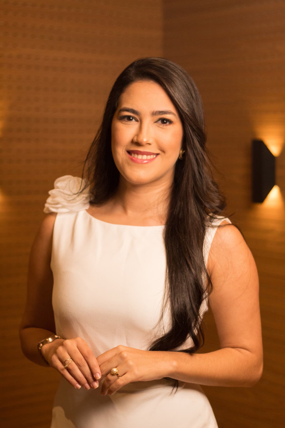 Lígia Ferreira sorrindo, de vestido branco, no escritório com painel de madeira e luz quente